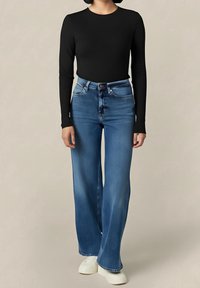 Černý top s dlouhým rukávem spojený s modrými širokými džíny s vysokým pasem, které mají zapínání na knoflíky a zip, a bílé obuv.