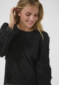 Sorte broderet bluse med teksturerede blomsterdetaljer, rund halsudskæring og lange ærmer med spidsdetaljer ved manchetterne.
