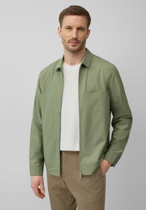 Lichtgroene ritsjack met een kraag, een enkele borstzak en een gladde textuur, gedragen over een witte gestructureerde shirt en tan broek.