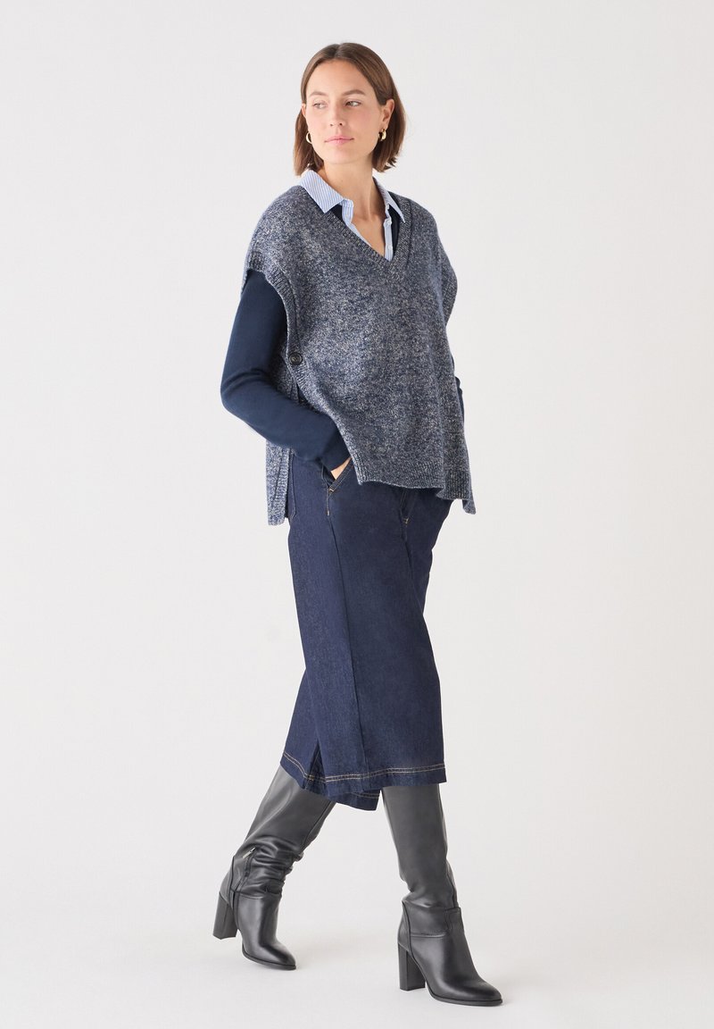 Blaue Strickponcho über einem marineblauen Langarmshirt und einem dunklen Jeansrock, kombiniert mit kniehohen schwarzen Stiefeln mit Absatz. Strukturiertes Material und geschichteter Stil.