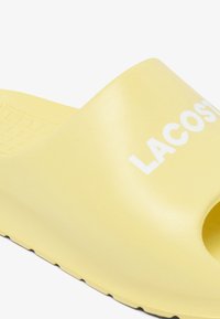 Sandale en caoutchouc jaune avec un bout ouvert, semelle intérieure texturée et logo "LACOSTE" blanc. Présente une finition lisse et une forme profilée.