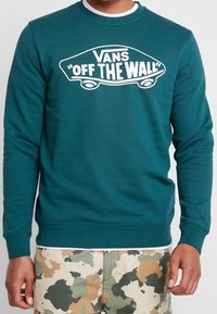 Sudadera verde azulado de algodón con mangas largas. Presenta un gran logo blanco "Vans 'Off the Wall'" en la parte frontal. Textura suave.