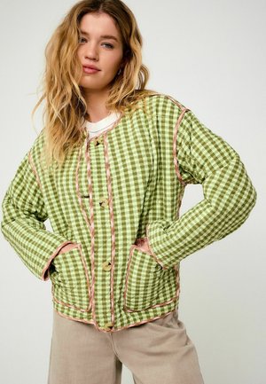 Young woman with loose blonde hair wearing beige pants and a green checkered quilted jacket with pink piping and front pockets.