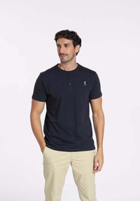 Polo en coton bleu marine avec un col rond, une patte de boutonnage à trois boutons et un petit logo sur le côté gauche. Associé à un pantalon beige.
