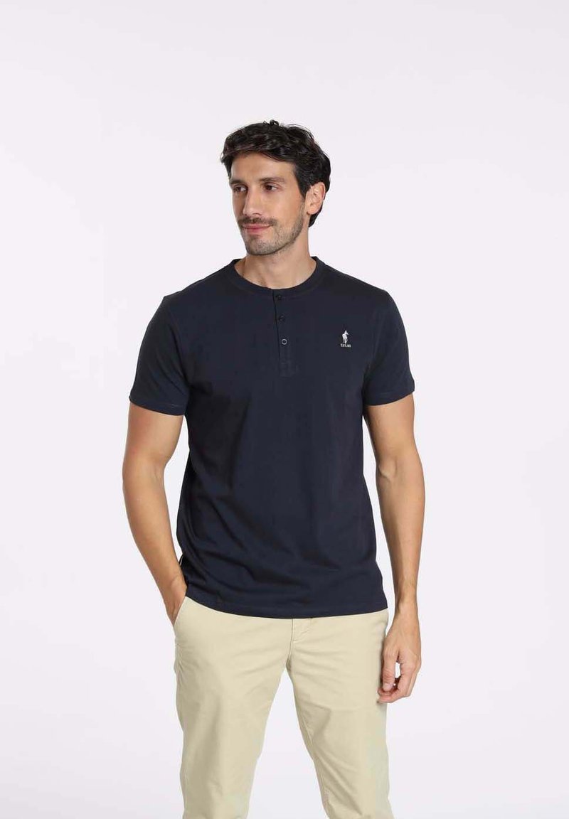 Polo en coton bleu marine avec un col rond, une patte de boutonnage à trois boutons et un petit logo sur le côté gauche. Associé à un pantalon beige.