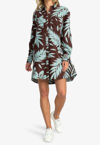 Robe chemise à manches longues en marron foncé avec des motifs de feuilles tropicales bleu clair, dotée d'une fermeture à boutons à l'avant et d'une coupe décontractée. Associée à des chaussures blanches.