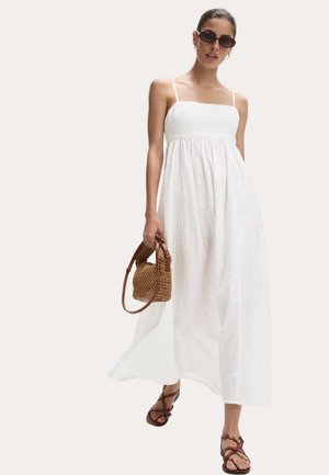 Robe maxi blanche avec un corsage smocké et des fines bretelles ; elle possède un tissu fluide et texturé. Associée à un sac à main tissé marron et des sandales.