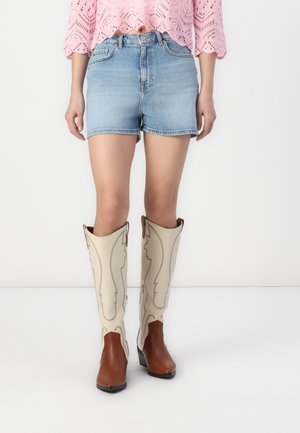 Bottes de cowboy beiges montant jusqu'aux genoux avec des accents marron et des coutures décoratives, portées avec un short en denim et un haut en crochet rose.