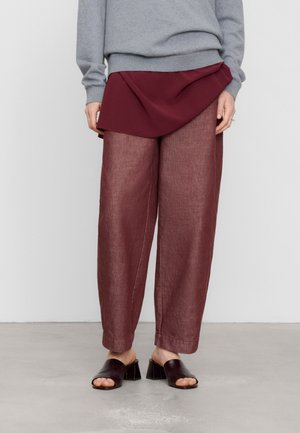 Moitié inférieure d'une personne portant un pantalon large bordeaux, un détail de jupe portefeuille assortie, un pull gris et des sandales noires à talon bloc ouvertes à l'avant.