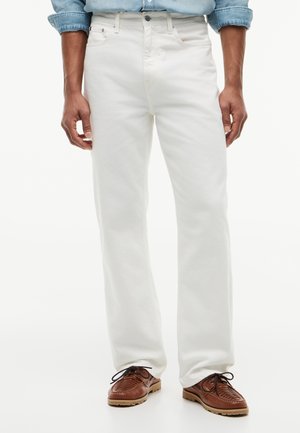 Mand iført hvide jeans, en lyseblå denimskjorte med op rullede ærmer og brune lædersko med snøre.