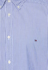 Camicia a righe azzurra e bianca con bottoni, dotata di un piccolo emblema sul lato sinistro del petto. Presenta un colletto a punta e bottoni bianchi.