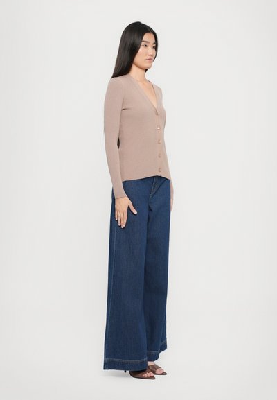 Cardigan beige a coste con scollo a V profondo e sette bottoni, abbinato a pantaloni in denim scuri a gamba larga e semplici décolleté open-toe.