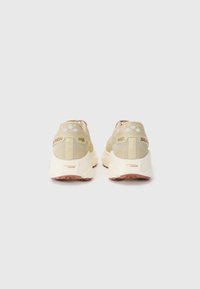 Beige atletik sko med mesh-overdel, tekstureret detaljering og gummisål. Har logo på hælen og tre cirkulære accenter på toppen.