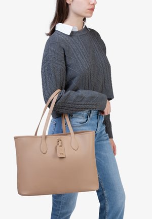 Borsa tote in pelle beige con texture liscia, doppio manico corto e ciondolo, portata da una persona che indossa un maglione di maglia grigia e jeans.
