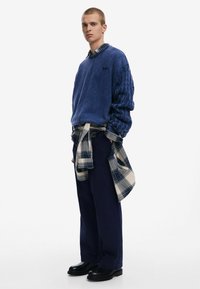 Blue fleece sweatshirt with textured sleeves, paired with dark navy pants. A plaid flannel is tied around the waist. Black boots complete the look.