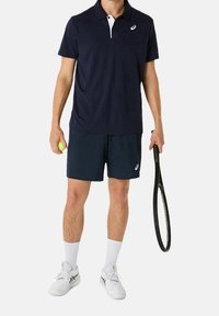 Man i marinblå tennisutrustning som håller en boll i ena handen och ett racket i den andra, klädd i vita sportskor och strumpor.