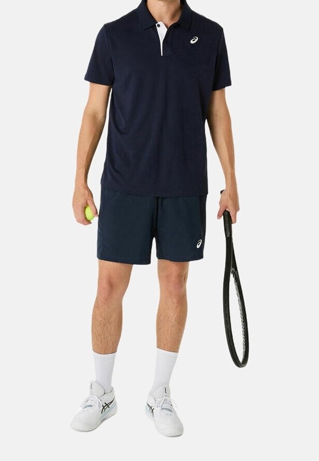 Man i marinblå tennisutrustning som håller en boll i ena handen och ett racket i den andra, klädd i vita sportskor och strumpor.