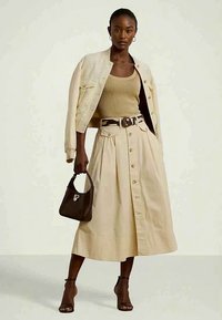 Nő bézs gombos szoknyát visel, vállán a hozzáillő zakóval, világosbarna bordás ujjatlan pólóval, sötétbarna táskával a kezében és fekete bokapántos magassarkú cipővel.