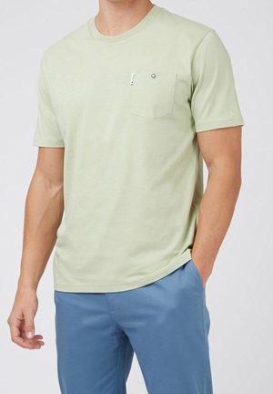 Mand iført lysegrøn kortærmet t-shirt med brystlomme og blå bukser, hånd i lommen mod ensfarvet baggrund.