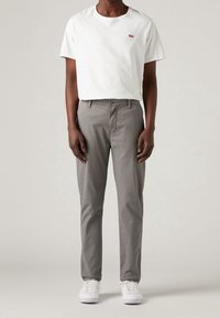 T-shirt bianco a girocollo con piccolo logo rosso, abbinato a pantaloni grigi chiari. Il modello indossa sneakers bianche, in piedi su uno sfondo neutro.