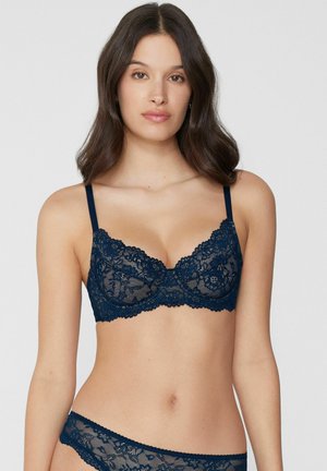 Jonge vrouw die een marineblauwe beha van bloemenkant en bijpassend slipje met lage taille draagt, staand tegen een effen lichte achtergrond.