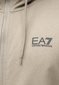 Μπεζ φούτερ με φερμουάρ, κατασκευασμένο από μαλακό ύφασμα, με κουκούλα με κορδόνι και σκούρο λογότυπο EA7 στο αριστερό μέρος του στήθους.