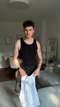 Jaunas vyras su tatuiruotėmis ir juoda beisbolo maike laiko dryžuotą marškinį šiuolaikiškame svetainėje su sofa ir valgomojo stalu.