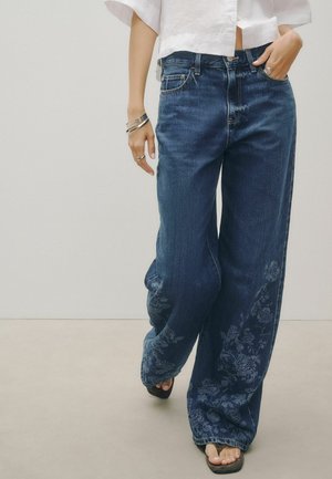Person trägt lockere dunkelblaue Jeans mit leichten Blumenmustern und ein weißes kurzes Hemd, barfuß in Sandalen, posiert mit einer Hand in der Tasche.