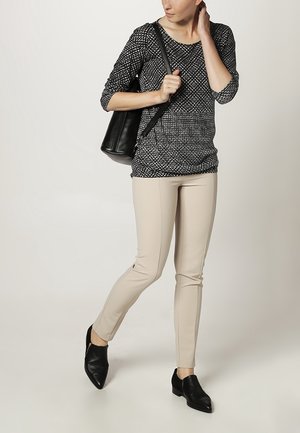 Femme portant un haut à motifs noir et blanc, un pantalon beige, des chaussures noires et un sac à bandoulière noir, debout devant un fond uni.