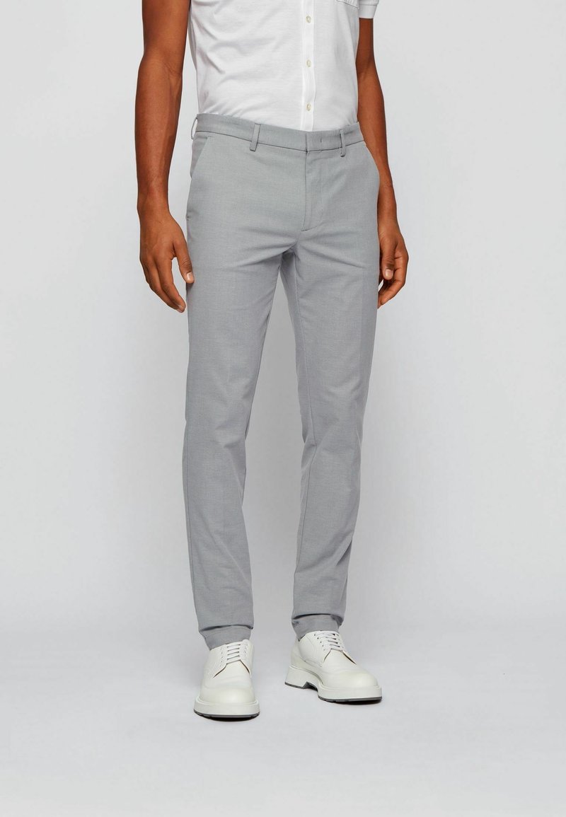 Homme portant un pantalon slim gris clair, une chemise blanche boutonnée et des chaussures blanches à lacets, debout sur un fond clair uni.