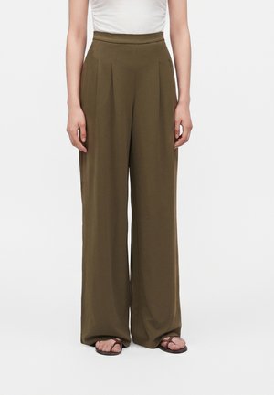 Persona che indossa pantaloni larghi verde oliva, top bianco e sandali marroni che sta in piedi su uno sfondo bianco semplice.