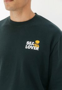 Hombre con sudadera verde oscuro con un pequeño pulpo amarillo y el texto "AMANTE DEL MAR" en el pecho sobre un fondo blanco.