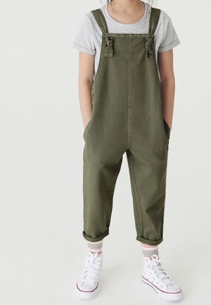 Enfant portant une salopette vert olive, un t-shirt gris à manches courtes, des chaussettes rayées et des baskets blanches montantes, debout les mains dans les poches.