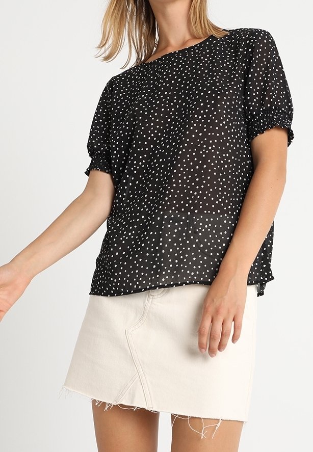 Černá průsvitná blůza s bílými puntíky, krátké nabírané rukávy, výstřih do půl kola, v kombinaci s bílou denimovou mini sukni s neupraveným lemem.