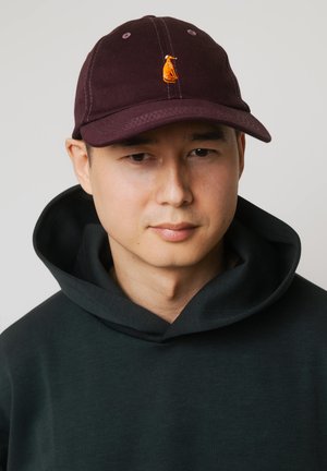 Mężczyzna w ciemnej bluzie z kapturem i ciemnoczerwonej czapce z małym pomarańczowym logo psa, na neutralnym tle, zwrócony twarzą do przodu z lekkim uśmiechem.