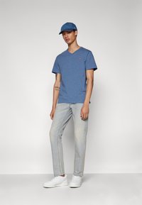 Blå V-hals T-skjorte, lyseblå denimjeans, hvite sneakers og en blå caps. Har et lite rødt logo på skjorten.