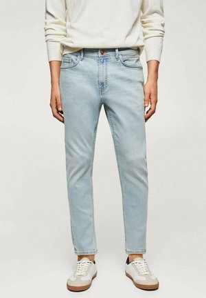 Mand iført lyseblå slim-fit jeans, hvid langærmet skjorte og hvide sneakers med brune såler mod en ensfarvet baggrund.