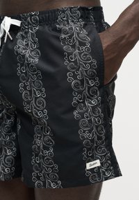 Gros plan d'une personne portant un short de bain noir avec des motifs de vigne blancs en spirale et un cordon blanc, main dans la poche latérale.