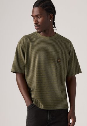 Homme aux cheveux tressés portant un t-shirt rayé vert et noir avec une poche poitrine, regardant vers le bas et à gauche, les mains dans les poches d’un pantalon noir.