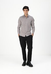 Chemise à motifs noir et blanc avec un pantalon noir. Le modèle se tient debout avec les mains dans les poches, portant des chaussures noires.