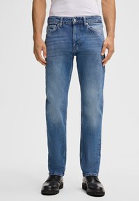 Jean da uomo in denim blu con leggera scoloritura, cinque tasche, vestibilità dritta e chiusura con bottone. Indossati con scarpe in pelle nere.