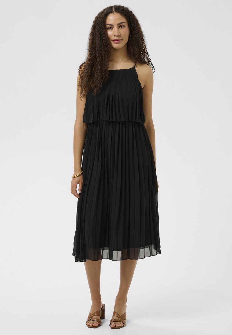 Femme aux cheveux bouclés portant une robe midi noire sans manches plissée et des sandales marron à talons sur un fond uni.