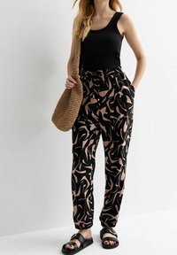 Vrouw in een zwart tanktop, zwarte en beige abstracte geblokte broek, zwarte sandalen en met een bruin geweven schoudertas.