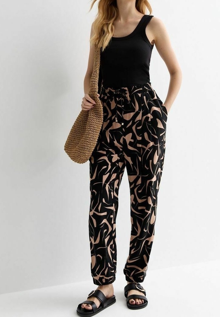 Vrouw in een zwart tanktop, zwarte en beige abstracte geblokte broek, zwarte sandalen en met een bruin geweven schoudertas.