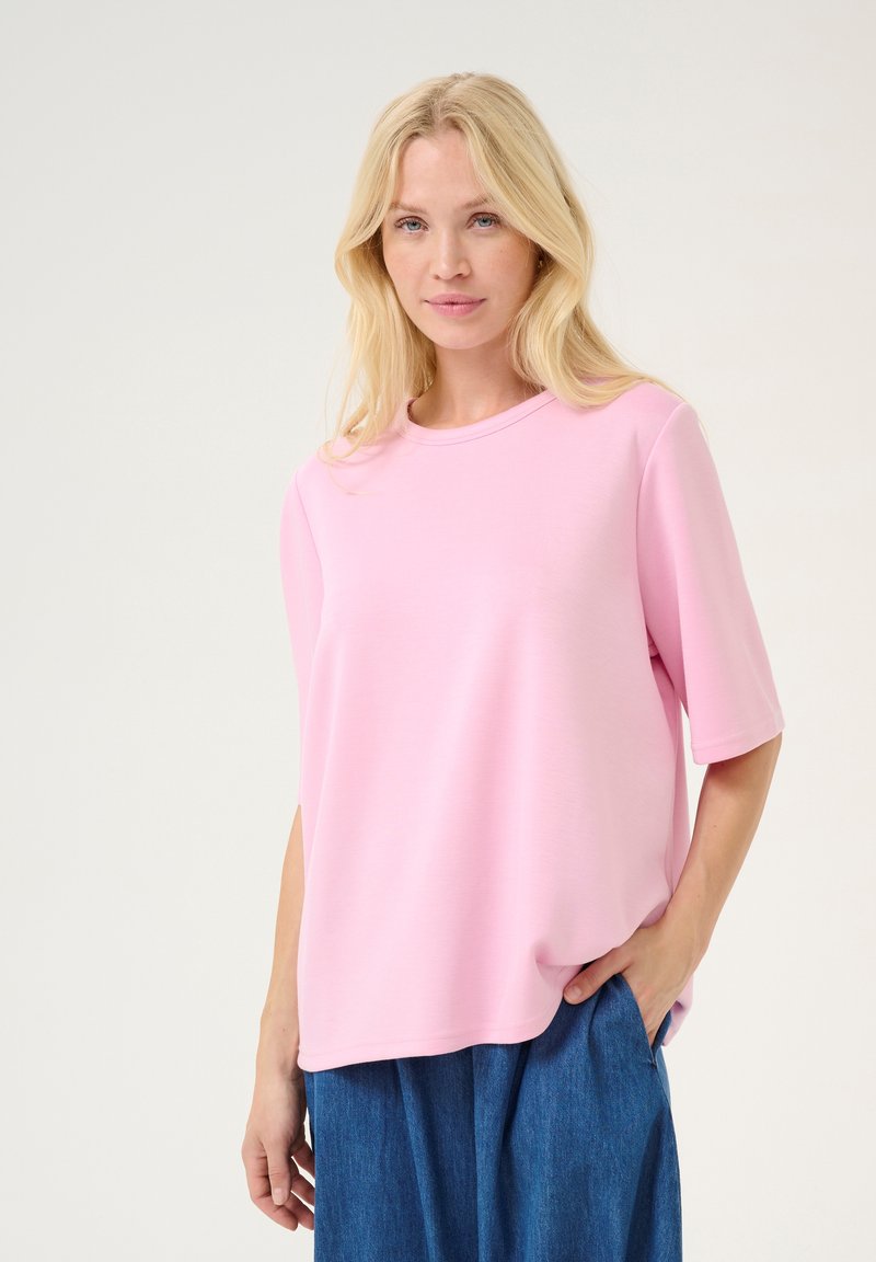 Roza kratka majica s kratkimi rokavi, izdelana iz gladkega materiala, sproščenega kroja in okroglega izreza, v kombinaciji z modrimi kavbojkami.