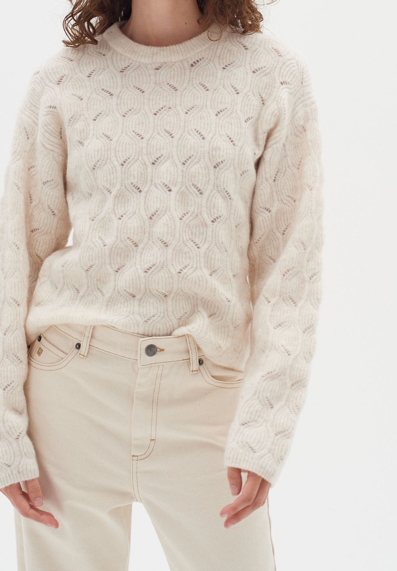 Beige stickad tröja med ett texturerat vågmönster, matchad med ljusbeiga hög midja jeans med framfickor och kontrasterande sömmar.