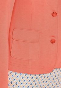 Koraalblazer met een patchzak, twee rode knopen en een soepele textuur; een gemusterde top verschijnt eronder met geometrische vormen in blauw en oranje.