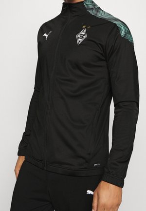 Chaqueta deportiva negra de Puma con el logo de Borussia Mönchengladbach, detalles verdes con patrón en los hombros y cremallera frontal, llevada por un modelo.