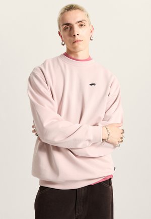 Rosa Sweatshirt aus weichem Stoff, mit Rundhalsausschnitt und kleinem schwarzen Skateboard-Logo. Über einem roten Shirt getragen.