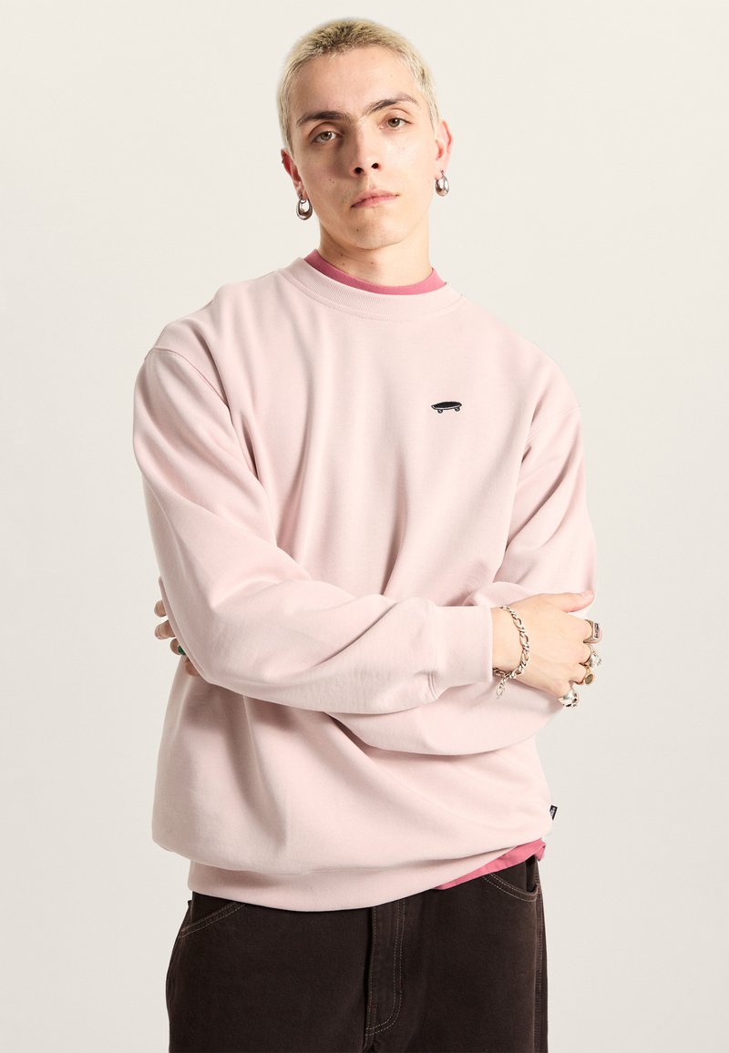 Lyserød sweatshirt lavet af blødt stof, med rund halsudskæring og et lille sort skateboardlogo. Laget over en rød skjorte.
